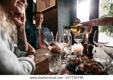 Man hand pouring white wine from the bottle into glasses with friends sitting around the table. Group of young people having food and drinks at restaurant.