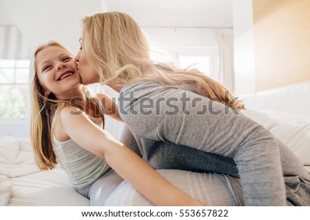 Young woman kissing little girl while sitting in bedroom. Loving mother and daughter on bed.