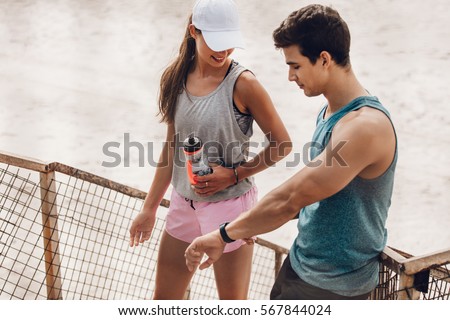 Young man and woman resting after running workout and checking fitness app on smart watch. Couple taking a break after workout outdoors and monitoring their performance on fitness device.