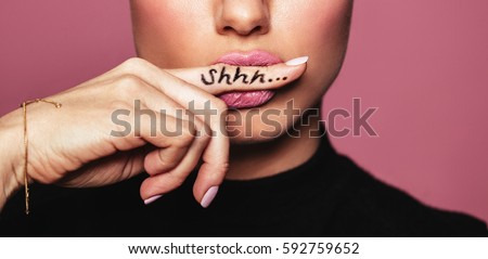 Cropped shot of young woman biting finger with shhh word. Young woman placing finger on lips asking shh against pink background.