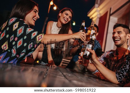 Group of friends toasting drinks at a party in evening. Young people hanging out at rooftop party and enjoying drinks.
