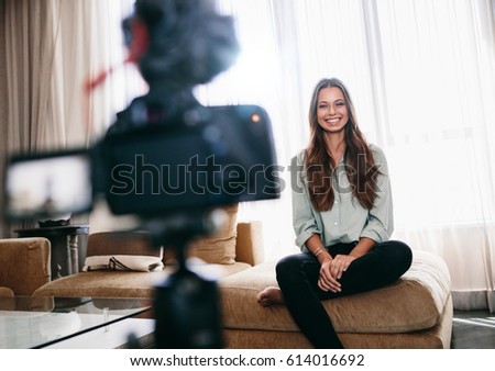 Young woman recording video on camera mounted on a tripod for her vlog. Pretty woman smiling at the camera sitting in her living room.