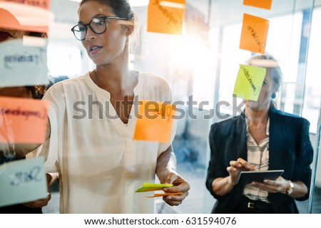 Portrait of creative professionals looking over a post it note wall and discussing. Female executives standing at the office behind glass wall with sticky notes.