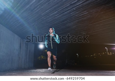 Full length shot of a young man jogging at night. Fit male athlete running under a bridge.