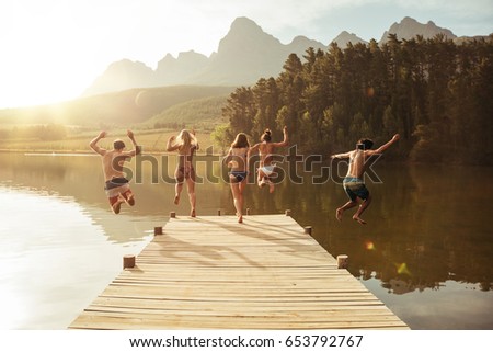 Group of young people jumping into the water from a jetty. Group of friends jumping from pier in the lake on a sunny day.