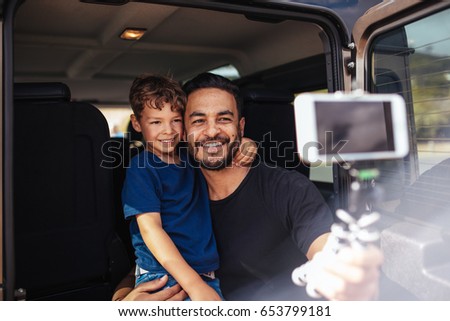 Handsome young dad and his cute little son sitting on back of the car making selfie using a smart phone. Father and son on road trip taking selfie.
