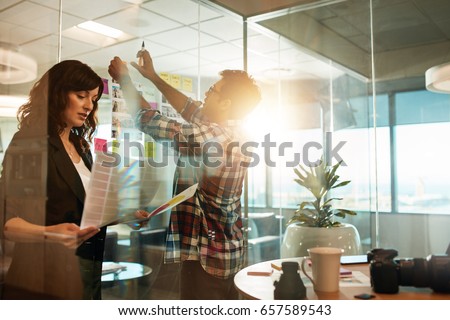Creative people working on new project. Woman looking at photographs and man sticking at contact sheet on glass wall in office.