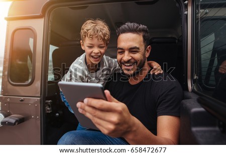 Shot of smiling father and son sitting at the back of the car with digital tablet. Young man and little boy having fun on road trip with digital tablet.