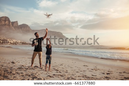 Full length shot of father and son operating the drone by remote control at the sea shore. Young man and little boy flying drone at the sea shore, having fun during summer vacation.
