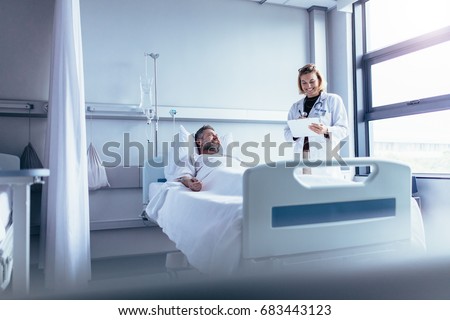 Female doctor talking to male patient in hospital bed. Smiling doctor with clipboard attending sick man in hospital ward.