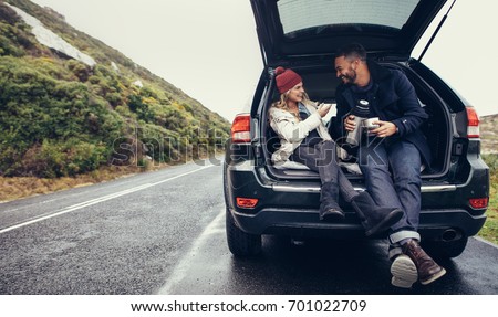 Happy young couple having a coffee break during road trip in countryside. Man and woman sitting in car trunk and having coffee.