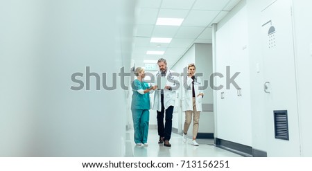 Group of medics with clipboard walking along hospital corridor. Doctor and nurse discussing medical report with female colleague talking on mobile phone.
