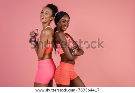Smiling fitness women standing back to back with water bottle. Fitness women relaxing after workout against pink background .