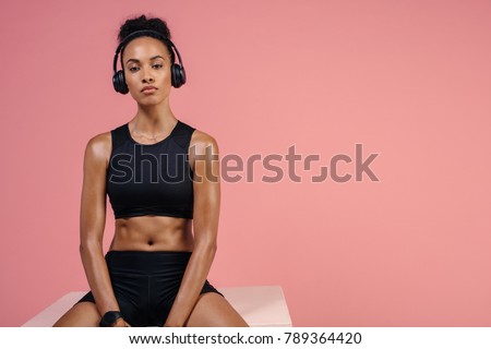 Strong young female athlete in sportswear and headphones sitting on box. African woman in sportswear relaxing after fitness training session.