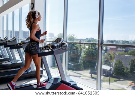 Woman running on treadmill at a panoramic window and listening to music via headphone at gym. Concept of healthy lifestyle.