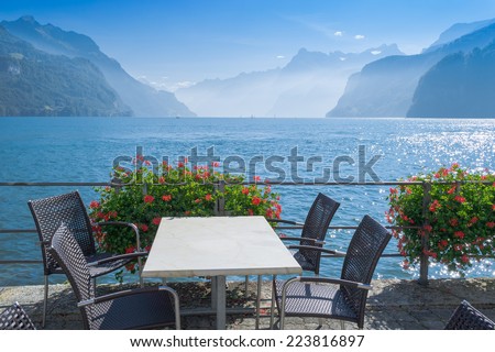 On the shore of  lake a small cafe, table and chairs. Flowers hanging on the railing. Silhouettes of  mountains in distance. Autumn day,  sun and  blue sky.