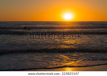 Sun set over the horizon. Pacific ocean and sky are painted in bright orange and gold colors. Waves beat against the sandy beach.