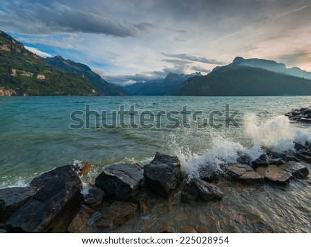 Storm on the lake. Splashing waves crashing on the shore rocks. Mountains on the horizon under cloudy sky