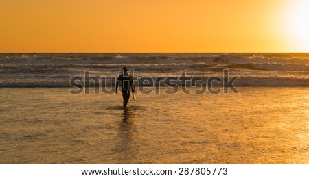 The male figure with a board for surf on a background of ocean waves. The setting sun paints the sky in bright orange, yellow, gold color