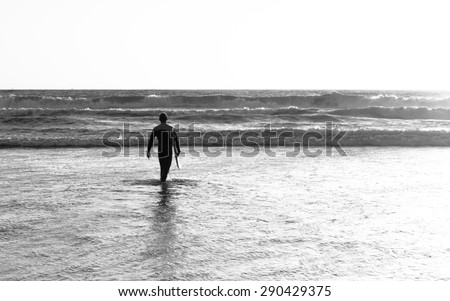 The male figure with a board for surf on a background of ocean waves. The setting sun paints the sky in bright orange, yellow, gold color. Vintage filter, monochrome. B&W