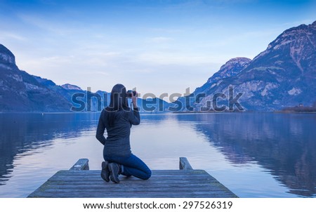 The female figure  is sitting with a camera in hand on a wooden pier. Mountain landscape at sunset, reflected in the lake. Clouds in the sky