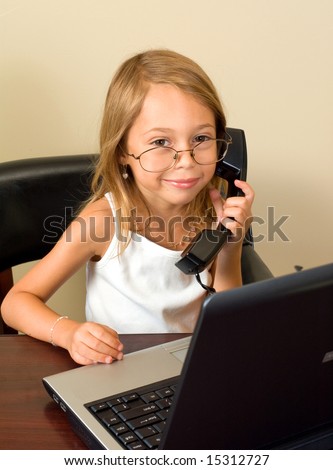 A young girl about 6 pretends to be busy at her notebook computer while wearing glasses too big for her head, talking on a telephone.