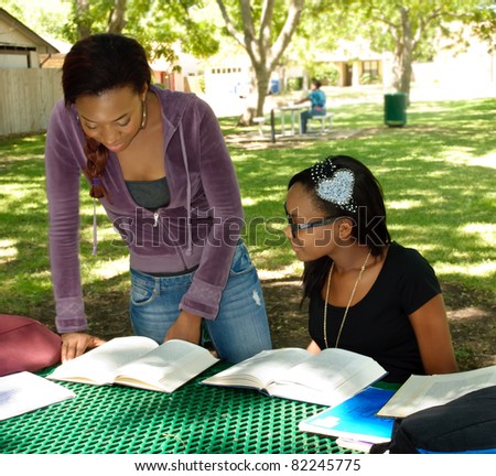 Two black teens study their books at the park, the older girls is standing and consulting a book.
