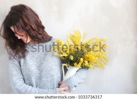 Young beautiful girl with flowers in hands, posing against concrete wall, minimalist style, mimosa in a flower pot