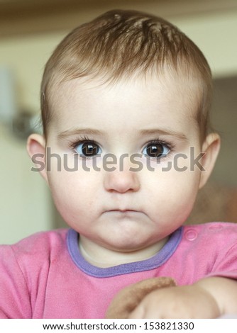 Baby girl, indoors, staring at camera, good expression, looking a bit bewildered/ grumpy. Head and shoulders shot. Wearing pink and lilac top. Big brown eyes.