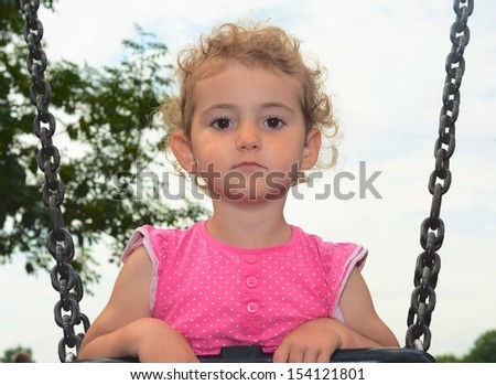 Young child on a swing at the park. Toddler girl smiling on a swing at the playground. She is wearing pink top and has blonde curly hair. Low viewpoint with white cloudy sky and trees background.
