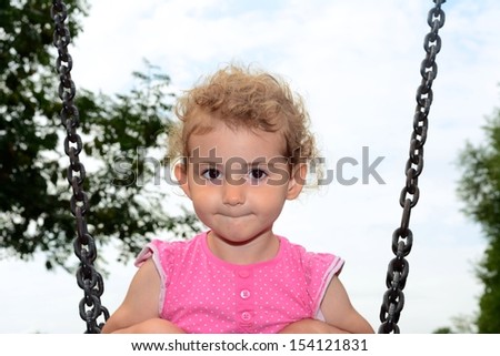 Young child on a swing at the park. Toddler girl smiling on a swing at the playground. She is wearing pink top and has blonde curly hair. Low viewpoint with white cloudy sky and trees background.