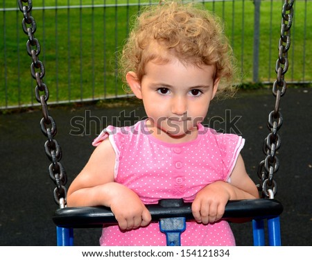 Young child on a swing at the park. Toddler girl smiling on a swing at the playground. She is wearing pink top, blue jeans and has blonde curly hair. Green grass backdrop.