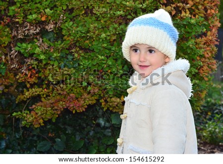Young child, girl, in woolly hat playing outside in the park on a cold day. She is wearing a white and blue hat and light coloured coat. She is smiling or laughing, having fun.