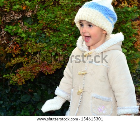 Young child, girl, in woolly hat playing outside in the park on a cold day. She is wearing a white and blue hat and light coloured coat. She is smiling or laughing, having fun.