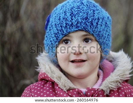 Young child, girl, with a bright blue woolly hat on. playing outdoors. She has beautiful big brown eyes and is playing in the woods.