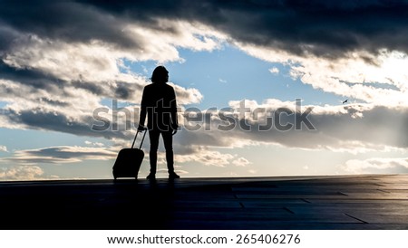 Man standing silhouetted to cloudy skies. Could be a traveler packed and ready with his trolley. Ready to travel with train or plain. Sunny skies in the background