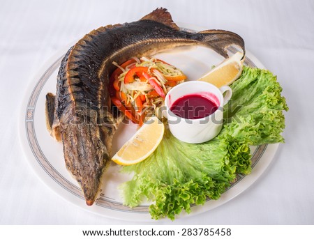 Baked sturgeon with vegetables and sweet red sauce. Plate located on a white canvas tablecloth background.