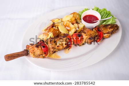 Grilled chicken shish kebab with roasted potatoes and delicious red sauce. Plate located on a white canvas tablecloth background.