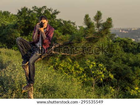 Young photographer with professional digital camera sitting on the tree and taking shot against park outdoors and city skylines.