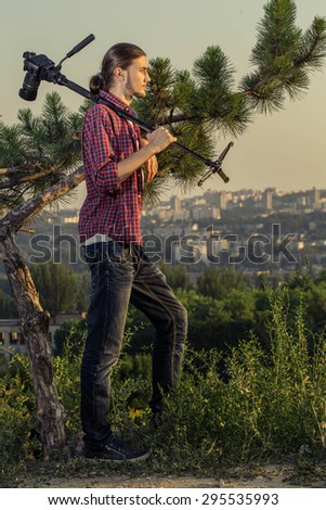 Young photographer with professional digital camera on monopod standing near the pine tree against blue sky and city skylines.