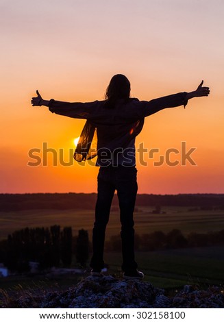 Silhouette of young longhair male model with thumbs-up hands at red and purple sunset sky.