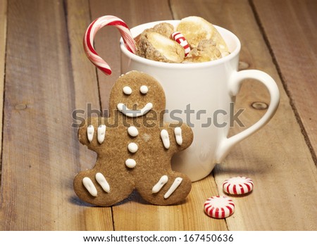 Mug of hot chocolate topped with toasted marshmallows and a candy cane with single gingerbread cookie leaning against mug and couple of round peppermint candies on the side on wooden table.