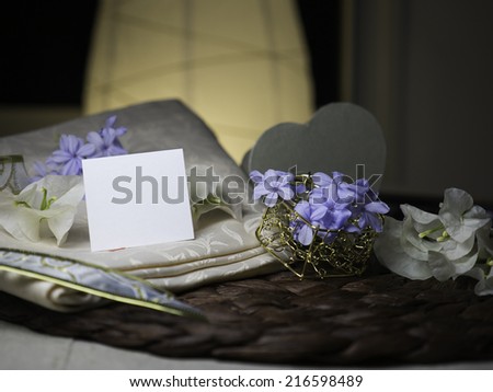 A blank note & white flowers laying on a nice textile near a golden heart box with blue flowers in it next to white flowers