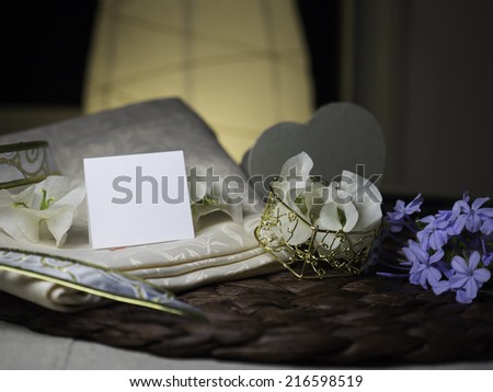 A blank note & white flowers laying on a nice textile near a golden heart box with white flowers in it next to blue flowers