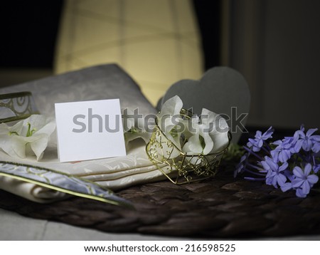 A blank note & white flowers laying on a nice textile near a golden heart box with white flowers in it next to blue flowers