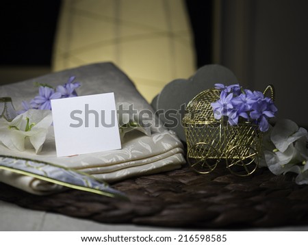 A blank note & white flowers laying on a nice textile near a golden stroller with blue flowers in it next to white flowers