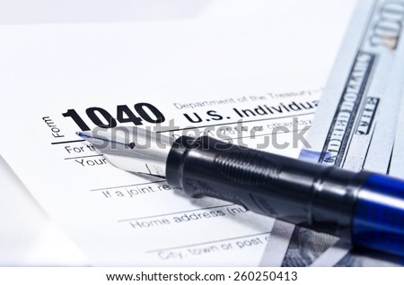 Tax form 1040 on a gray background, blue pen, and 100 dollar bills out of focus