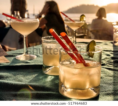 Margarita cocktail glasses on a beach bar table with people, sunset, sea and mountains background