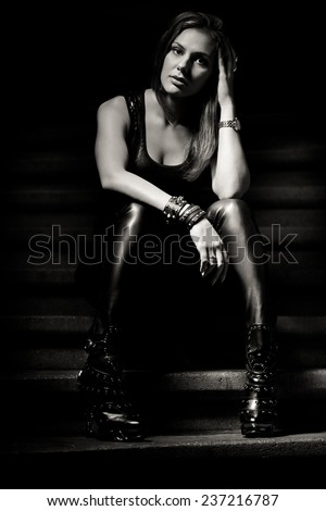 Beautiful sexy metal rock and roll woman in leather outfit sitting on the stairs. Street photo of urban metal rock woman fan. Sexy vamp femme fatale sensual edgy lady