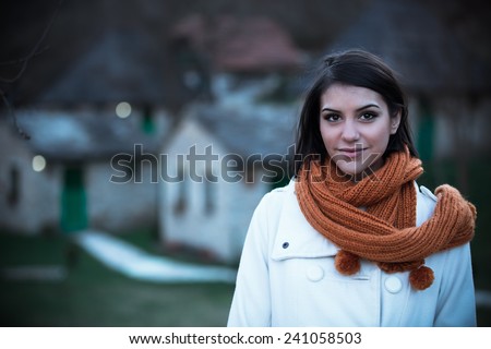 Beautiful girl in the winter coat standing near the winter resort,mountain chalets in the evening.December without snow.Tourist spending winter holidays at cottage in the mountains